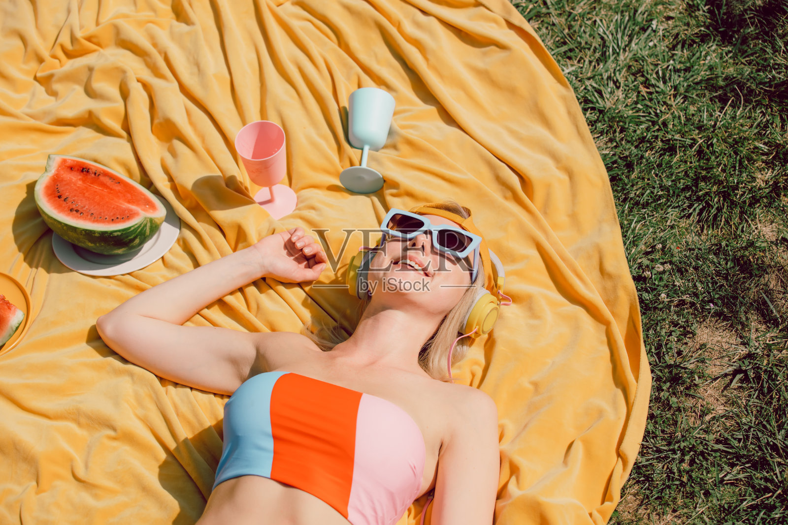 戴着墨镜和耳机的时尚女子躺在布上，旁边是绿草地上的西瓜照片摄影图片