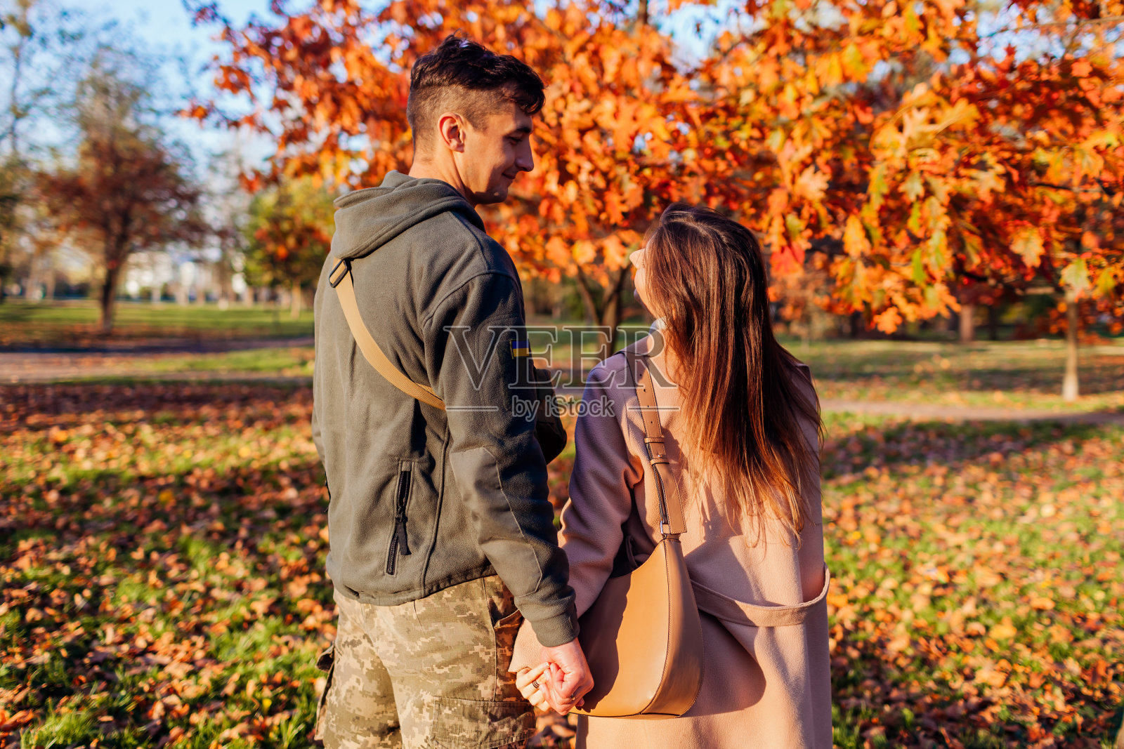 身穿乌克兰制服的士兵和他的妻子在秋天公园散步的背影。久别重逢的情侣拥抱。照片摄影图片