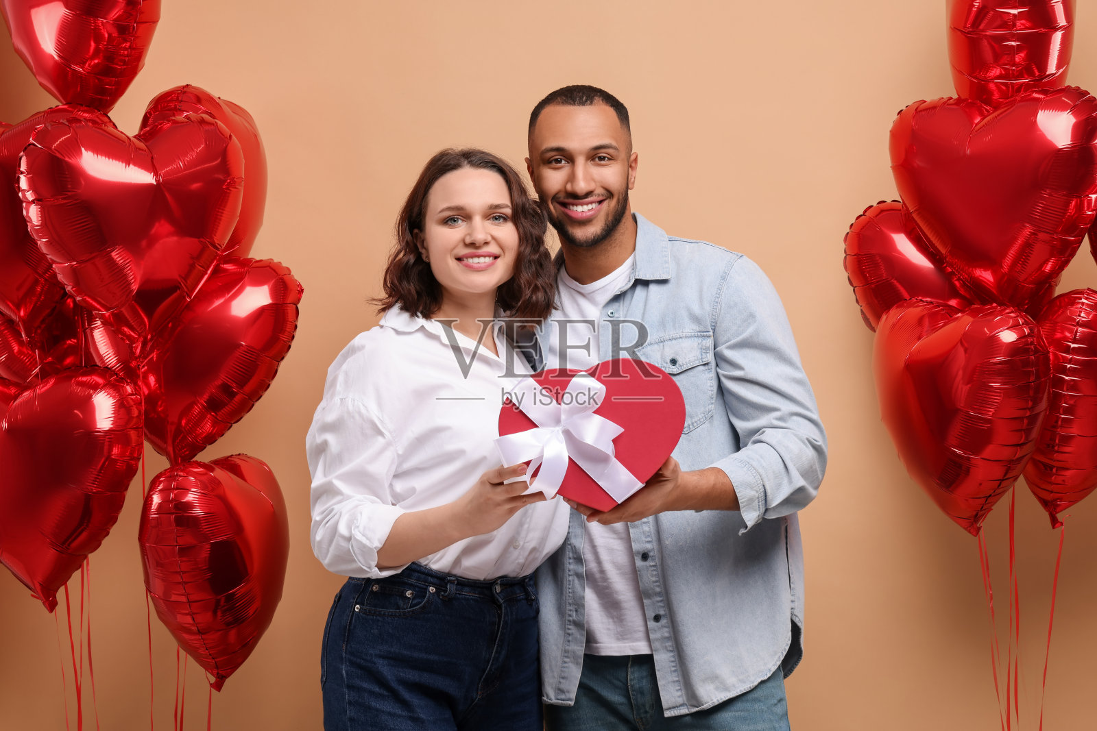 可爱的夫妇与礼品盒附近心形气球米色背景。情人节庆祝活动照片摄影图片