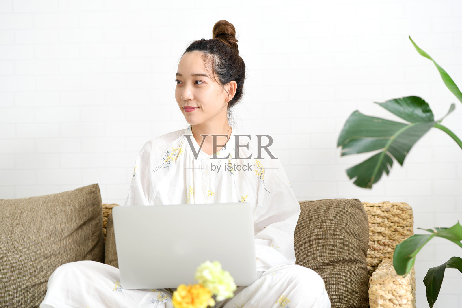 一个穿着睡衣的年轻亚洲女子坐在沙发上，微笑着操作笔记本电脑的场景。照片摄影图片