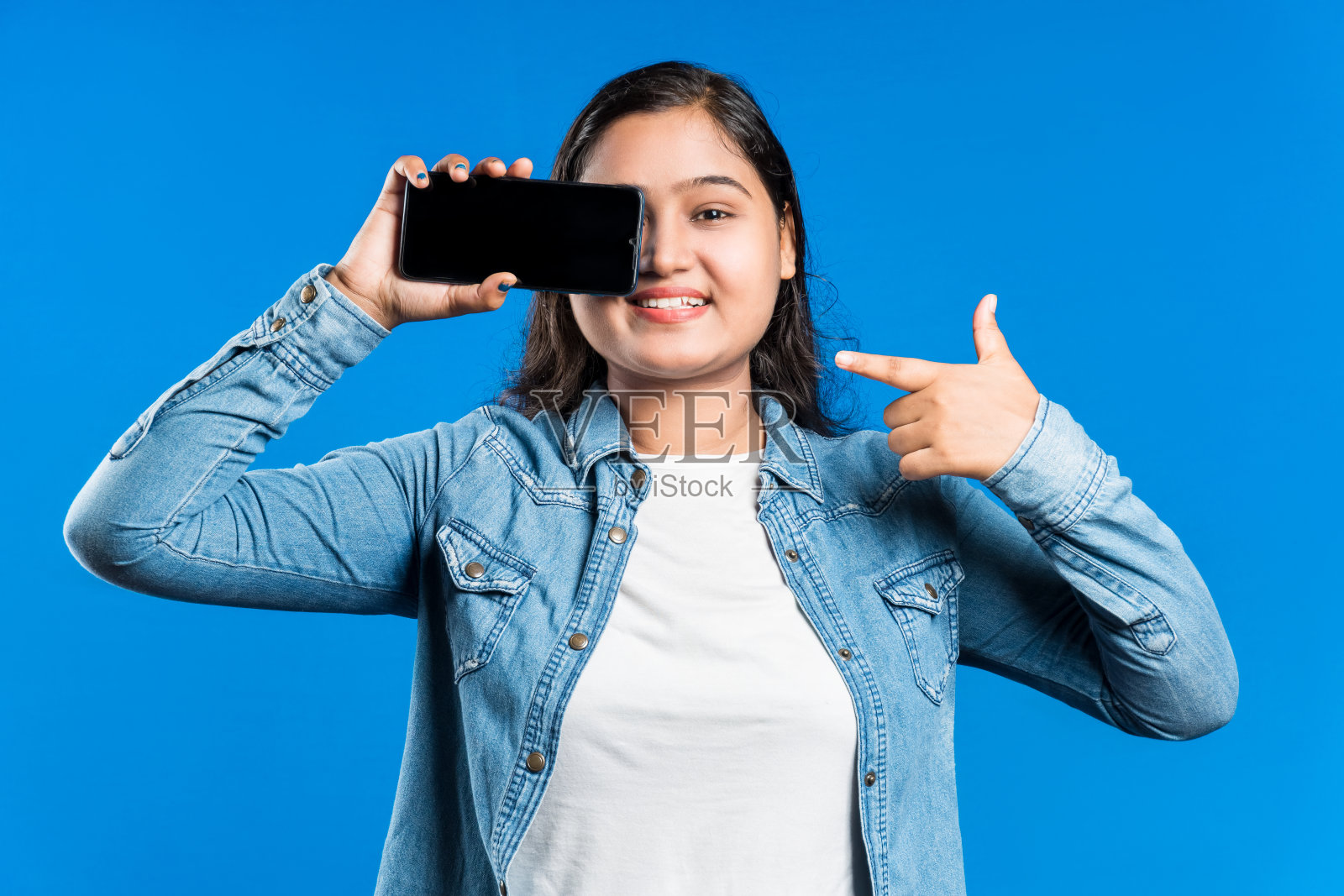 快乐的亚洲女人用智能手机遮住眼睛，用手指指着它照片摄影图片