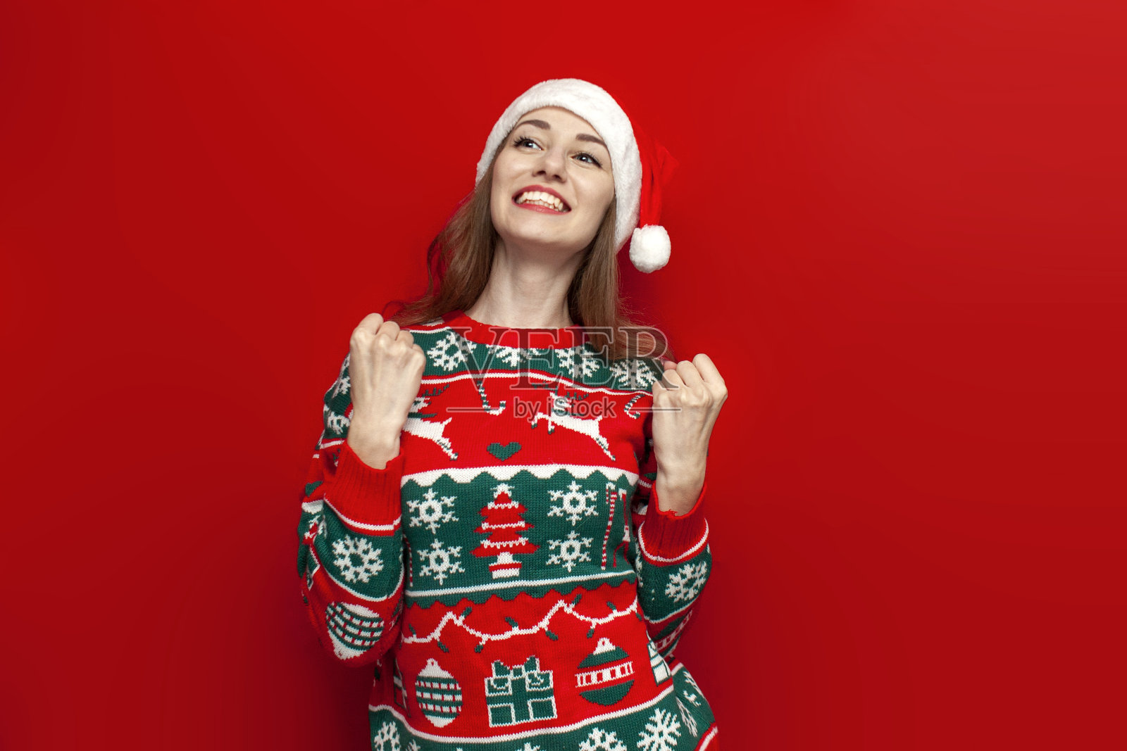 年轻可爱的女孩穿着圣诞毛衣，戴着圣诞帽，庆祝胜利，并在红色背景上大喊，胜利的女人照片摄影图片
