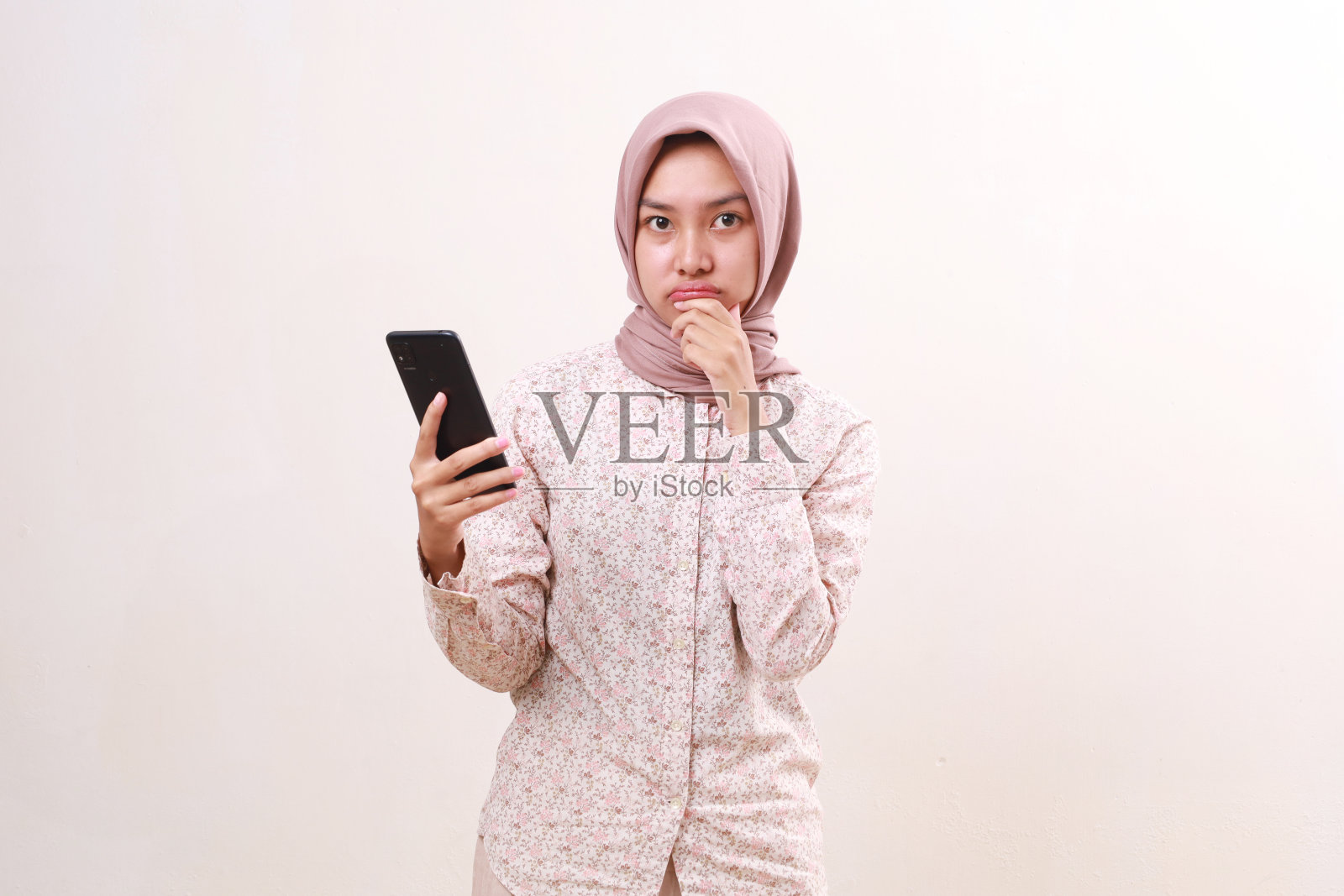 困惑的年轻亚洲穆斯林女孩拿着手机站着。孤立于白色照片摄影图片