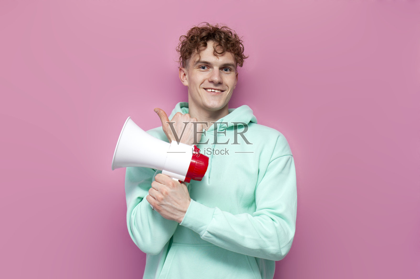 穿着薄荷色连帽衫的年轻卷发男子拿着扩音器，在粉红色的孤立背景上宣布新闻照片摄影图片