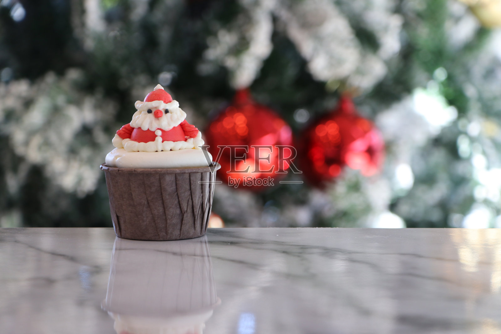 用奶油装饰的纸杯蛋糕。这是一个棕色杯子里的圣诞老人图案。放置在圣诞树的焦点背景与白色的雪和两个红色的球。照片摄影图片