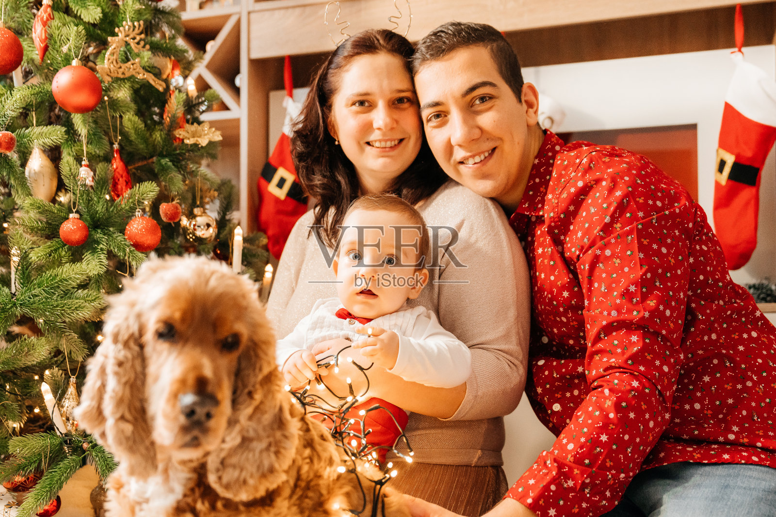 宝宝穿着圣诞服装，拿着礼物站在圣诞树旁。小男孩庆祝圣诞节。新年假期。年轻的家庭妈妈，爸爸和小儿子在圣诞树的背景。冬季新年的概念。照片摄影图片