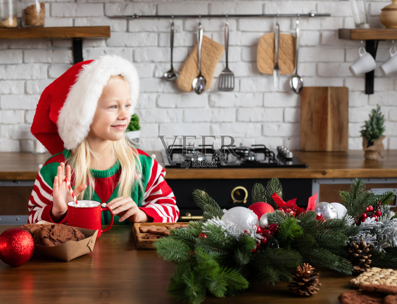 孩子打扮成小精灵拿着拐杖糖笑。孩子坐在厨房的木桌旁，红杯子里盛着热巧克力和棉花糖照片摄影图片