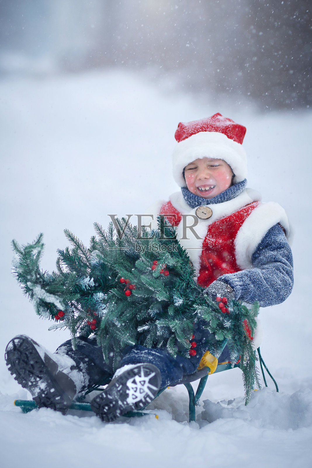 一个孩子穿着新年服装，在大雪中给圣诞老人举着一棵圣诞树。照片摄影图片