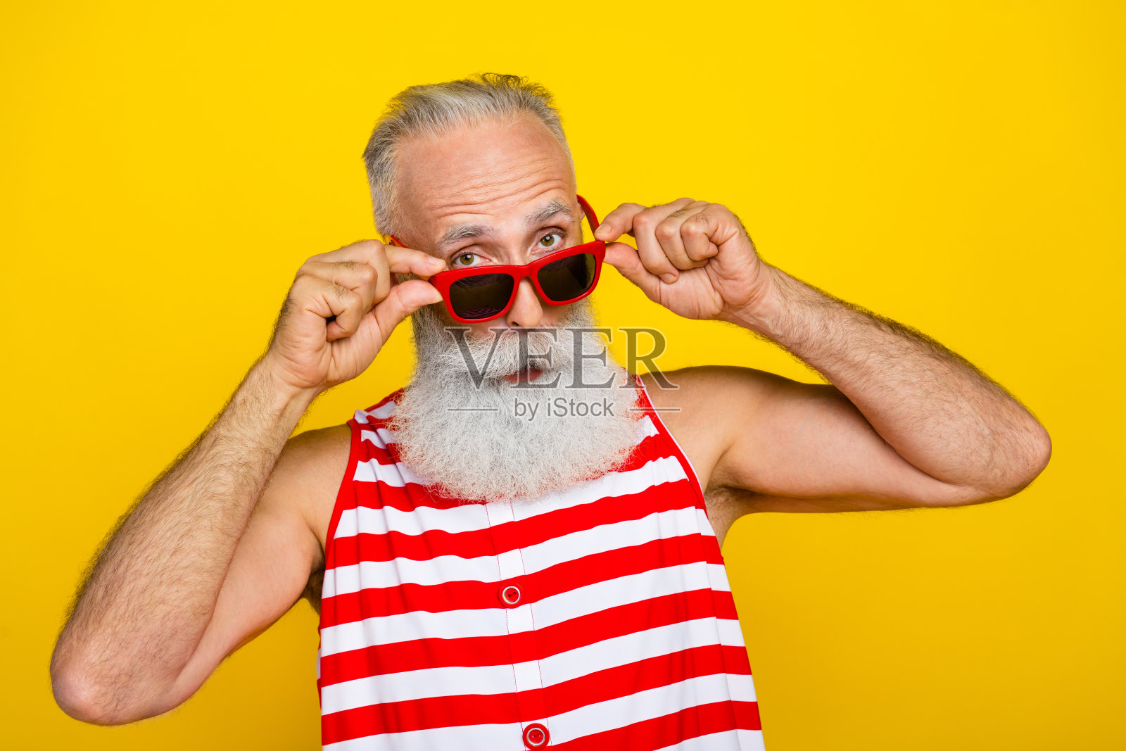 照片上开朗多疑的老年男子身穿红色条纹泳衣，双手手臂戴着墨镜望着你空旷孤立的黄色背景照片摄影图片
