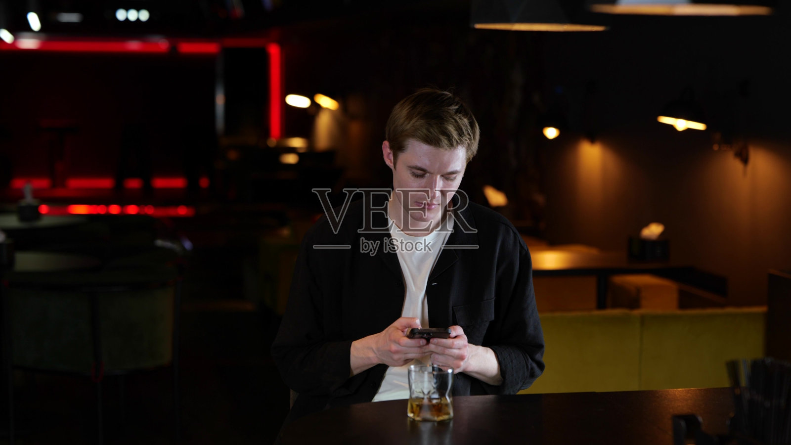 心烦意乱的年轻人一边看短信，一边拿着手机，喝着威士忌，坐在餐厅里。生活方式的概念。慢动作照片摄影图片