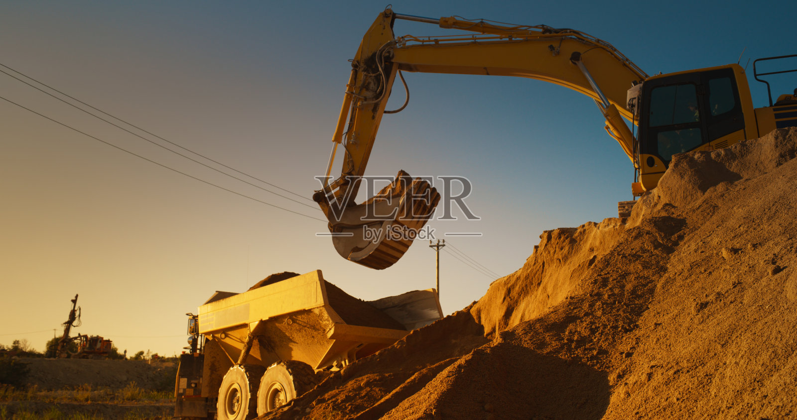 阳光明媚的晚上，建筑工地:工业挖掘机将沙子装进卡车。建设新公寓楼的过程。工人操作重型机械完成新的房地产项目。照片摄影图片