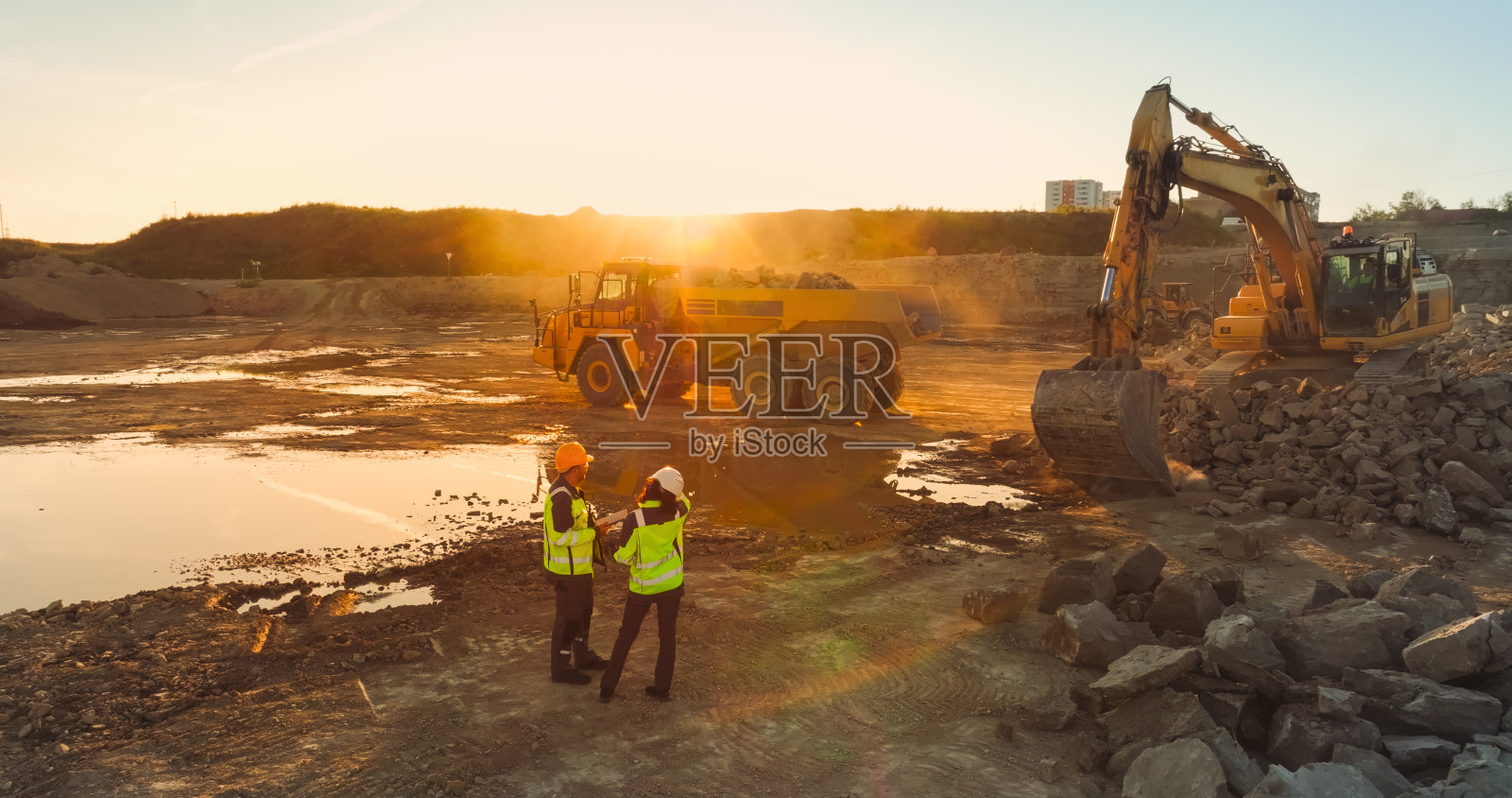 空中无人机拍摄的建筑工地在阳光明媚的晚上:工业挖掘机挖掘岩石，并把它们装上卡车。土木工程师和建筑师观察过程，讨论房地产项目照片摄影图片