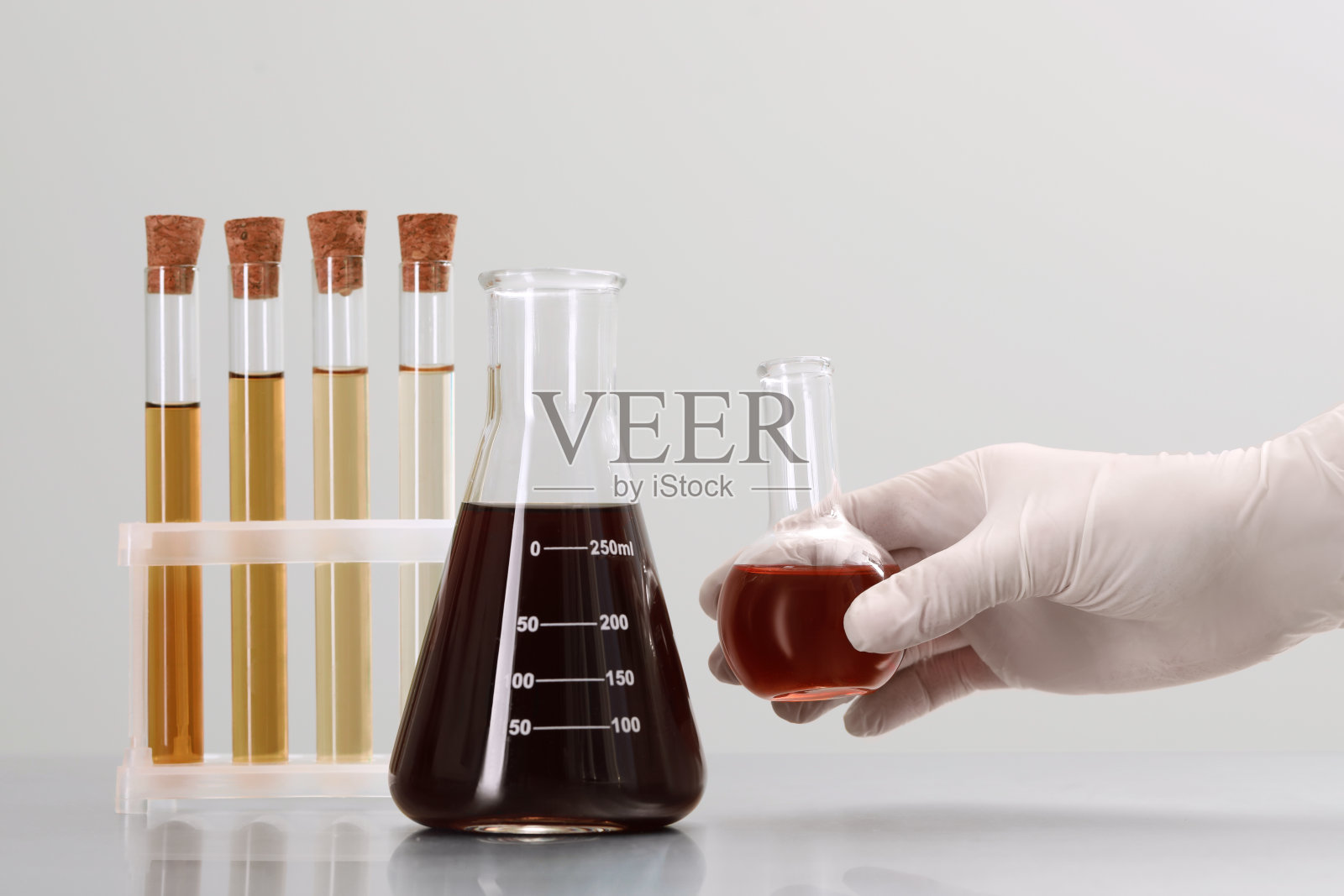 科学家拿着装有棕色液体的圆底烧瓶靠近浅色背景的实验室玻璃器皿，特写照片摄影图片