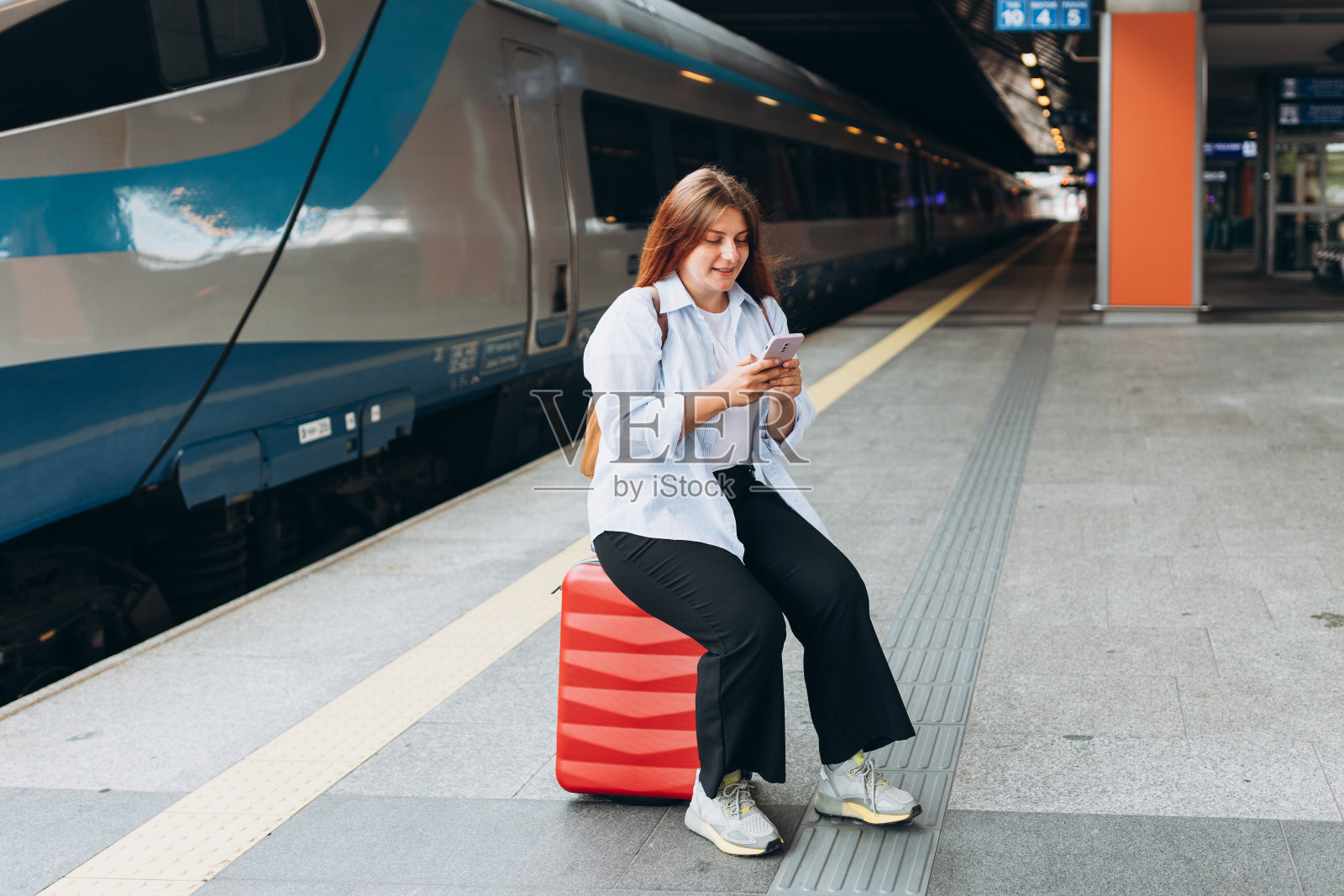 年轻的红发女子背着背包拿着智能手机等火车。铁路运输概念，妇女提着箱子走在火车站站台上。坐火车去度假，全身都是照片摄影图片