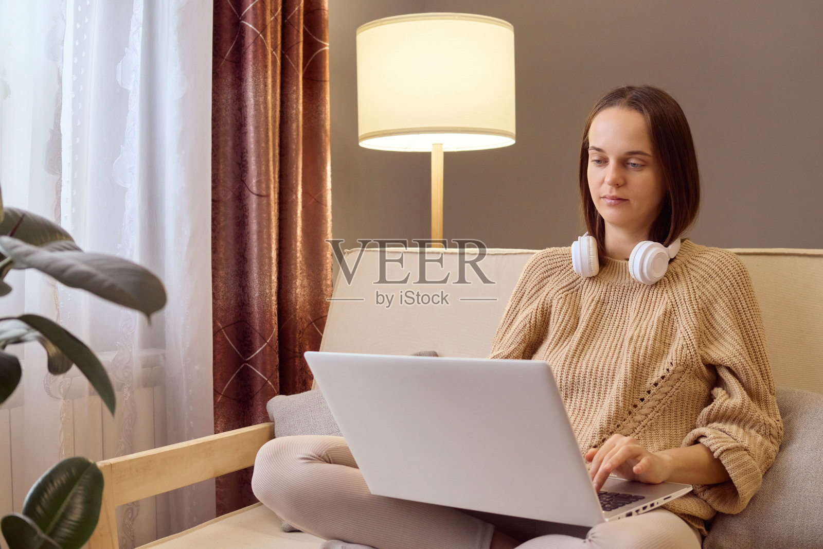 穿着针织毛衣的棕色头发的女人坐在家里的沙发上使用笔记本电脑漂亮的女孩购物或在社交网络上聊天看电影在电脑项目上工作的自由职业者。照片摄影图片