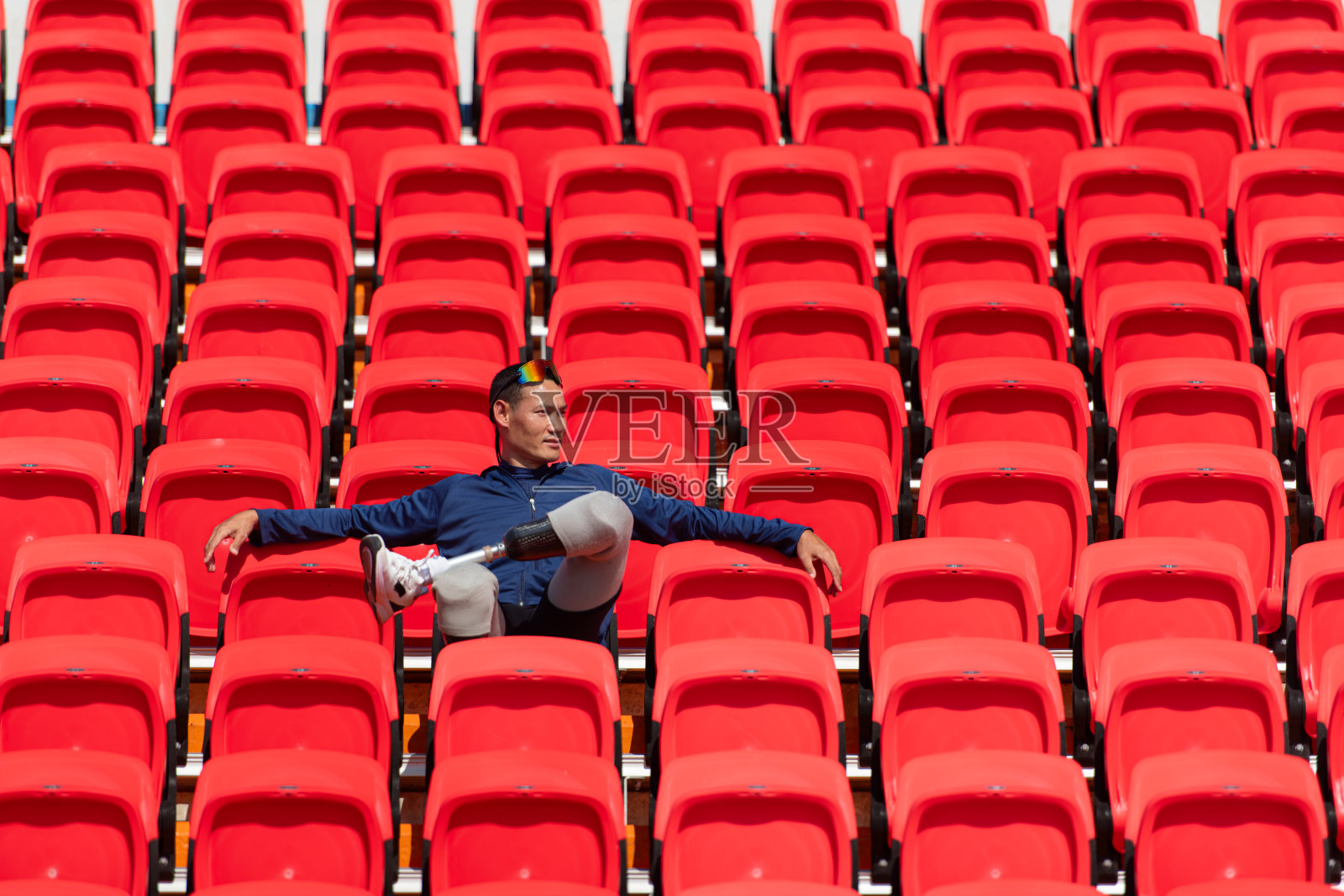 身穿蓝色运动衫的残疾运动员坐在体育场的红色座位上，为跑步训练做准备。照片摄影图片