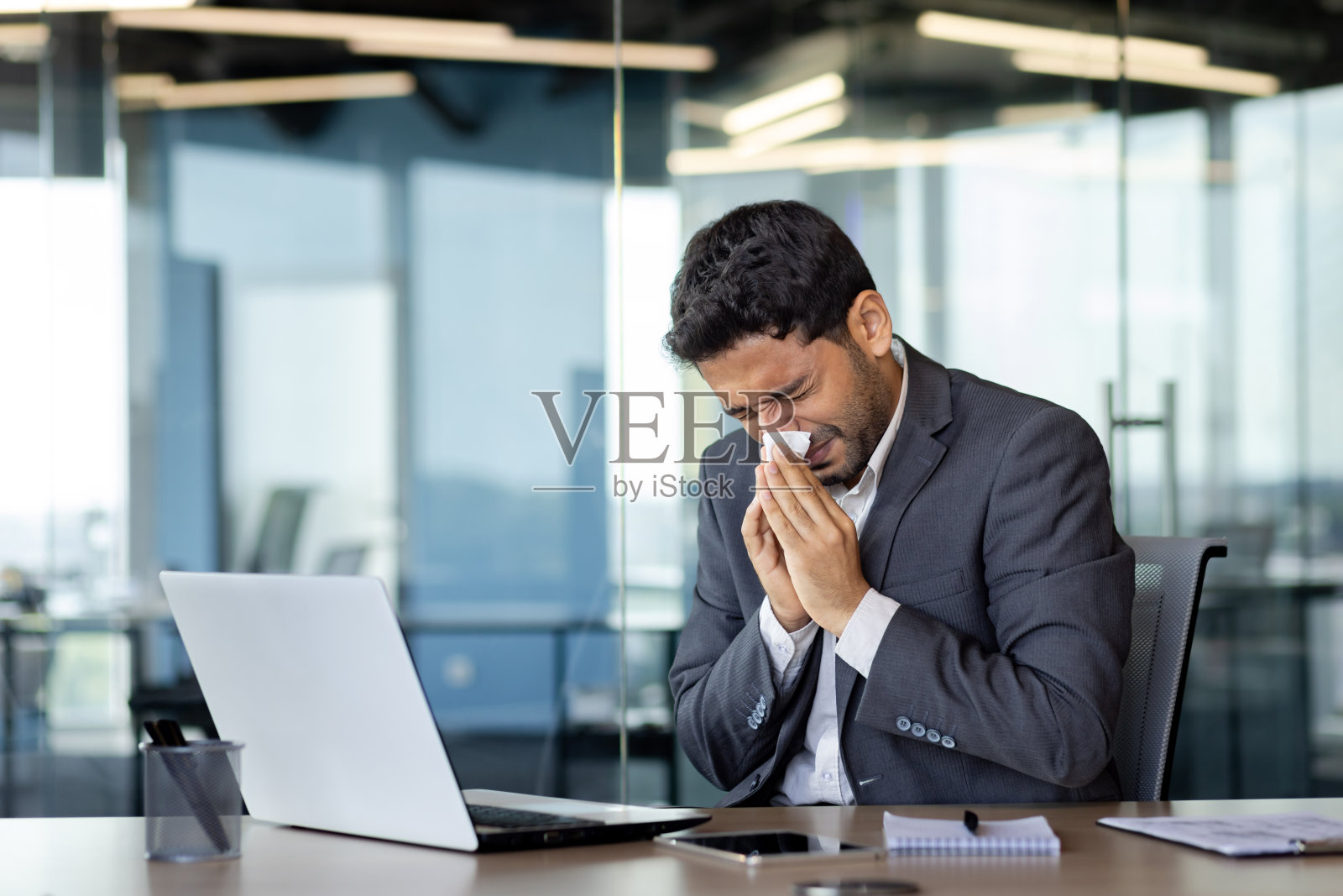 在办公室工作场所，一名男子对着纸巾打喷嚏，一名商人因流鼻涕而生病，在工作场所因过敏而工作，使用笔记本电脑工作照片摄影图片