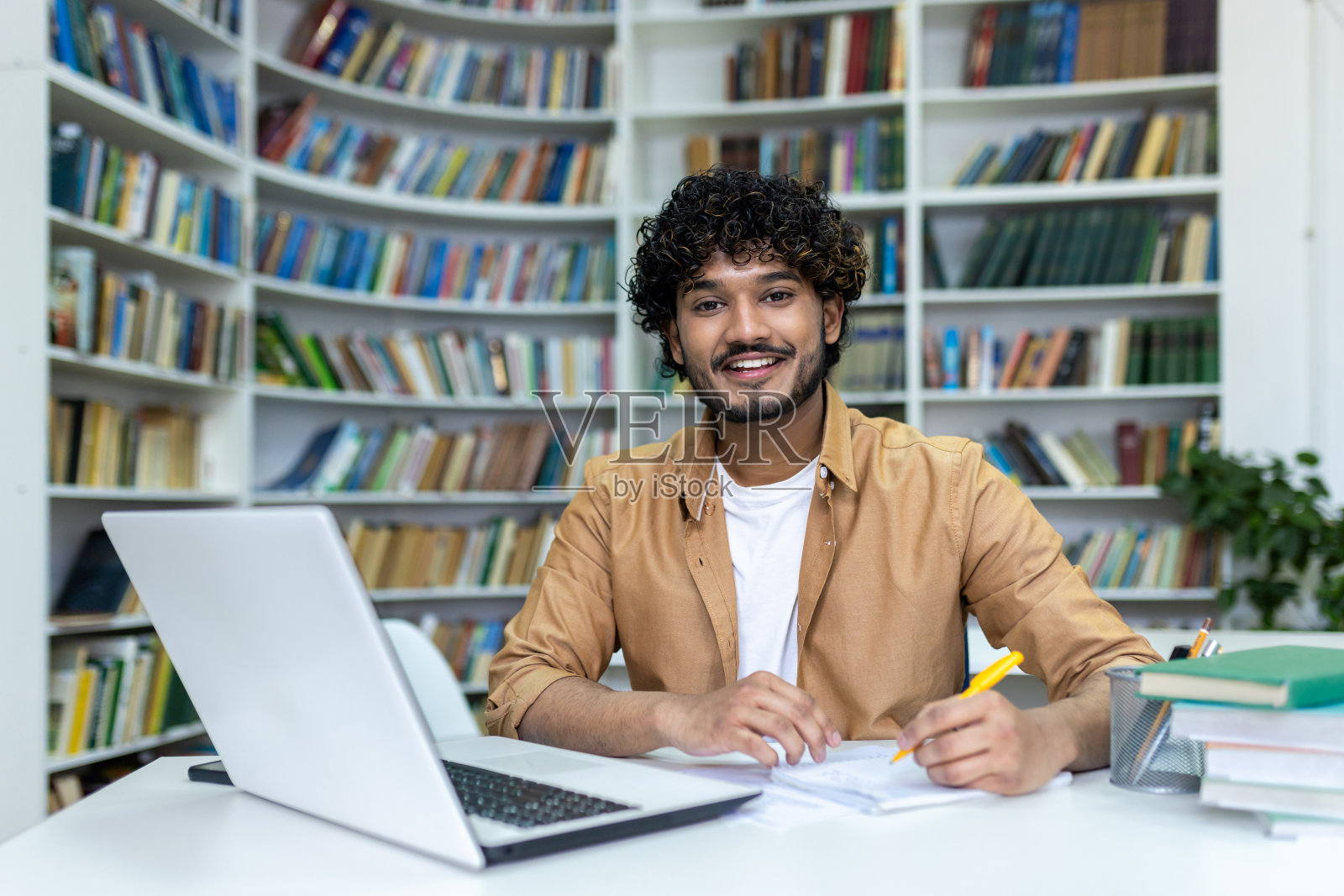 一个年轻的印度男学生在校园图书馆学习，用笔记本电脑在笔记本上写下讲课内容。他微笑着看着镜头照片摄影图片