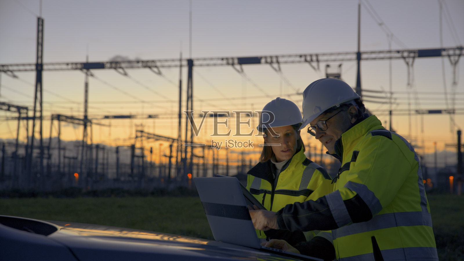 日落时分，电站汽车引擎盖上，男工程师在笔记本电脑上向女同事解释策略照片摄影图片