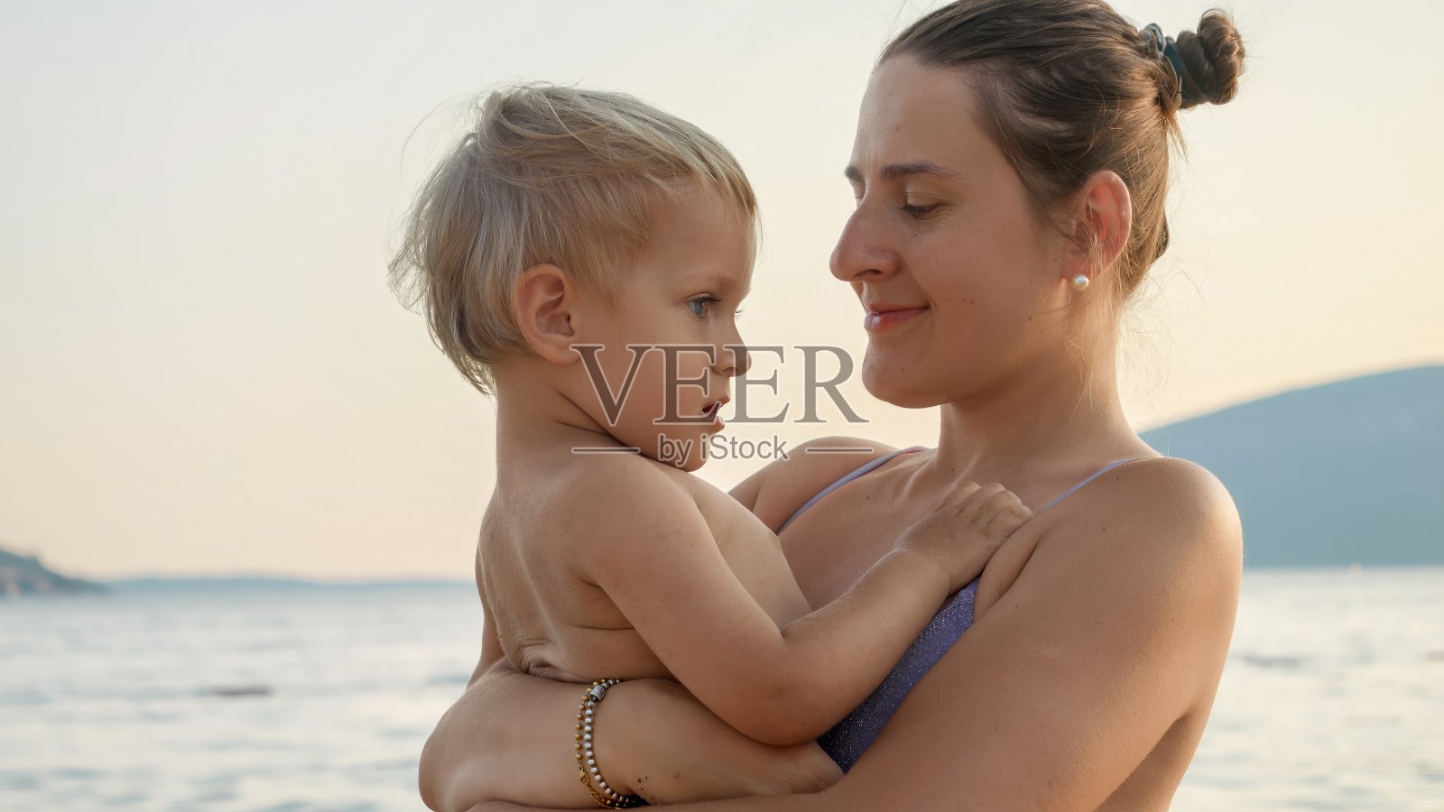 夕阳下的沙滩上，穿着比基尼的微笑女子抱着她的儿子。假期、暑假、旅游照片摄影图片