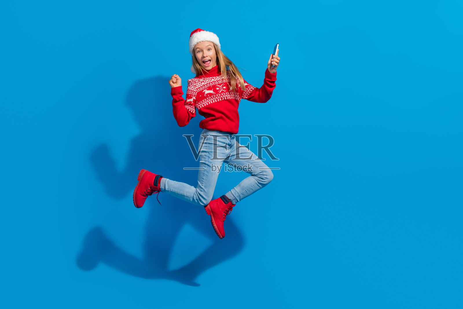 大喜的女学生穿着红色毛衣，圣诞帽飞扬，拿着智能手机，在蓝色背景下的圣诞节照片摄影图片