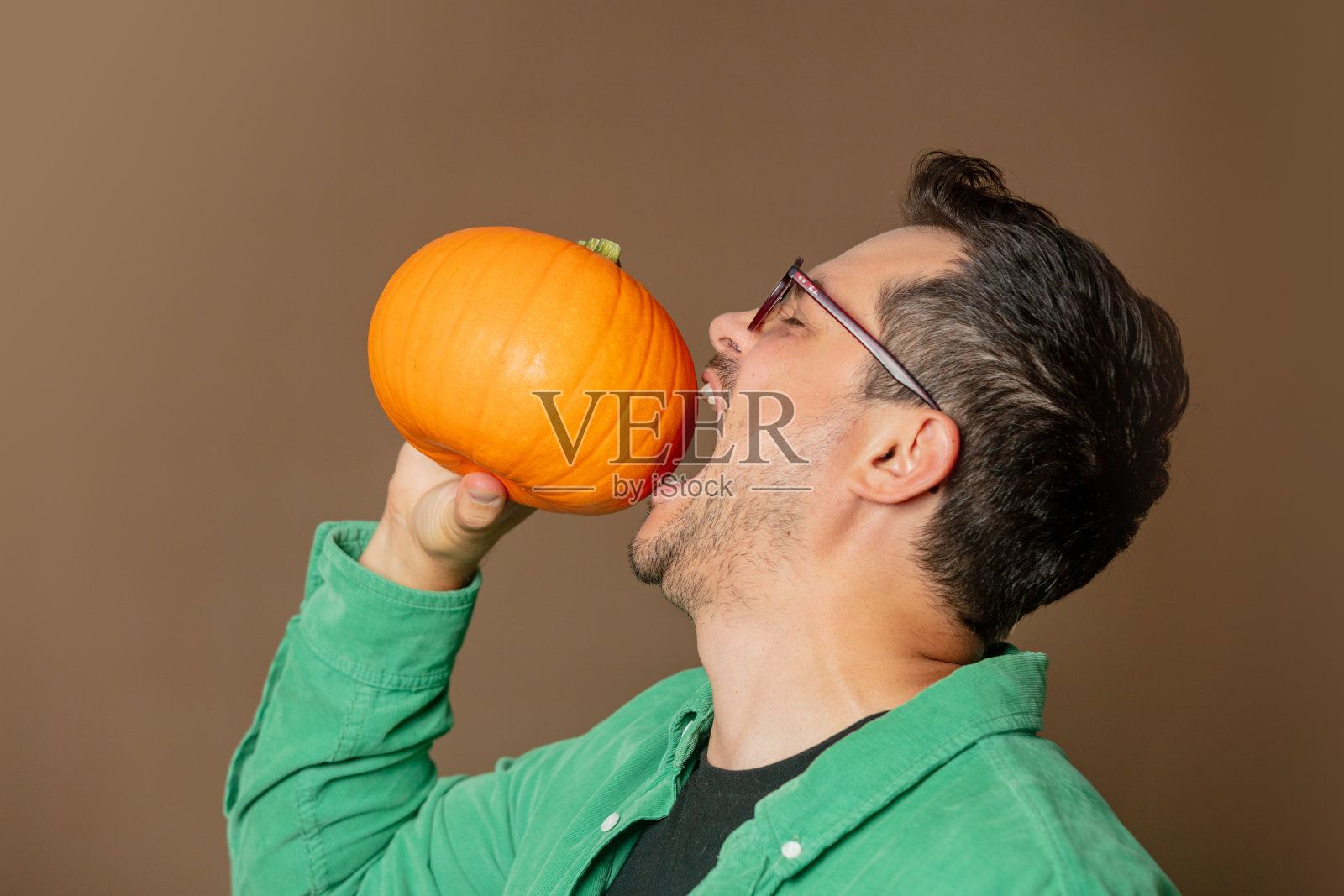 一个戴着眼镜、穿着绿色衬衫的时髦男人手里拿着南瓜，背景是棕色的照片摄影图片