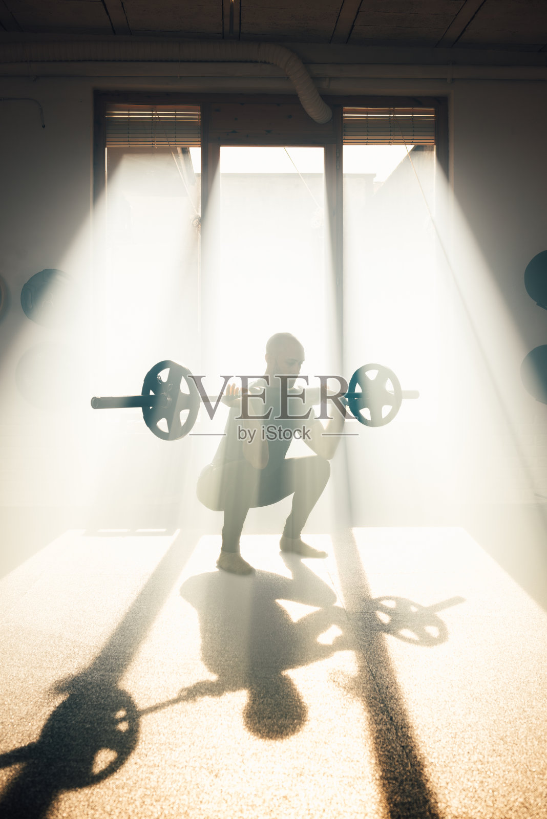 阳光下健身房里，一个强壮的男人举着杠铃蹲着照片摄影图片