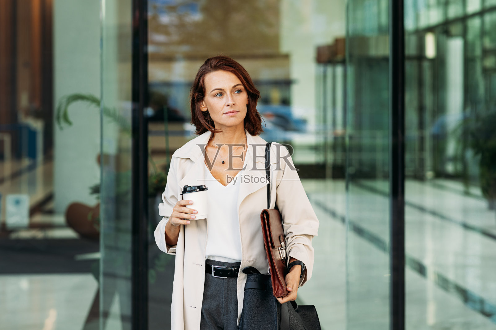 中年女商人端着咖啡从商业大楼走出来。在商业大楼里，一头姜黄色头发的自信女商人。照片摄影图片
