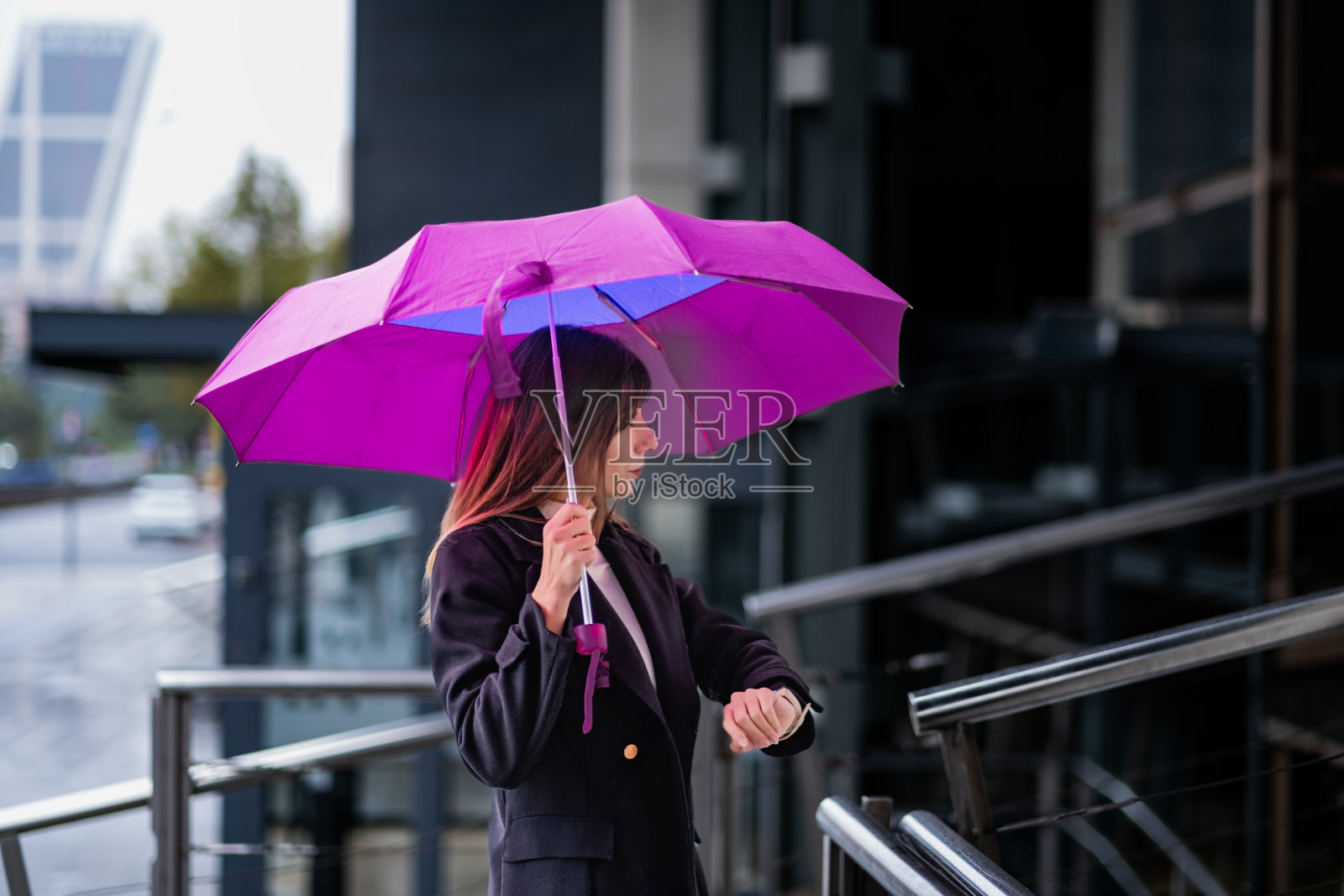 时髦的黑发女人，雨天的马德里，拿着雨伞，穿着黑夹克，等待着，玻璃建筑。照片摄影图片