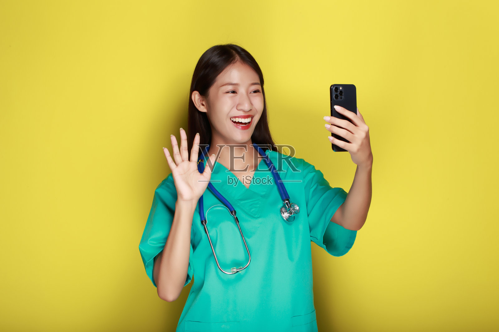 一个美丽的年轻女子在黄色背景下的肖像，亚洲妇女穿着医生的制服，戴着听诊器，拿着手机摆姿势。照片摄影图片