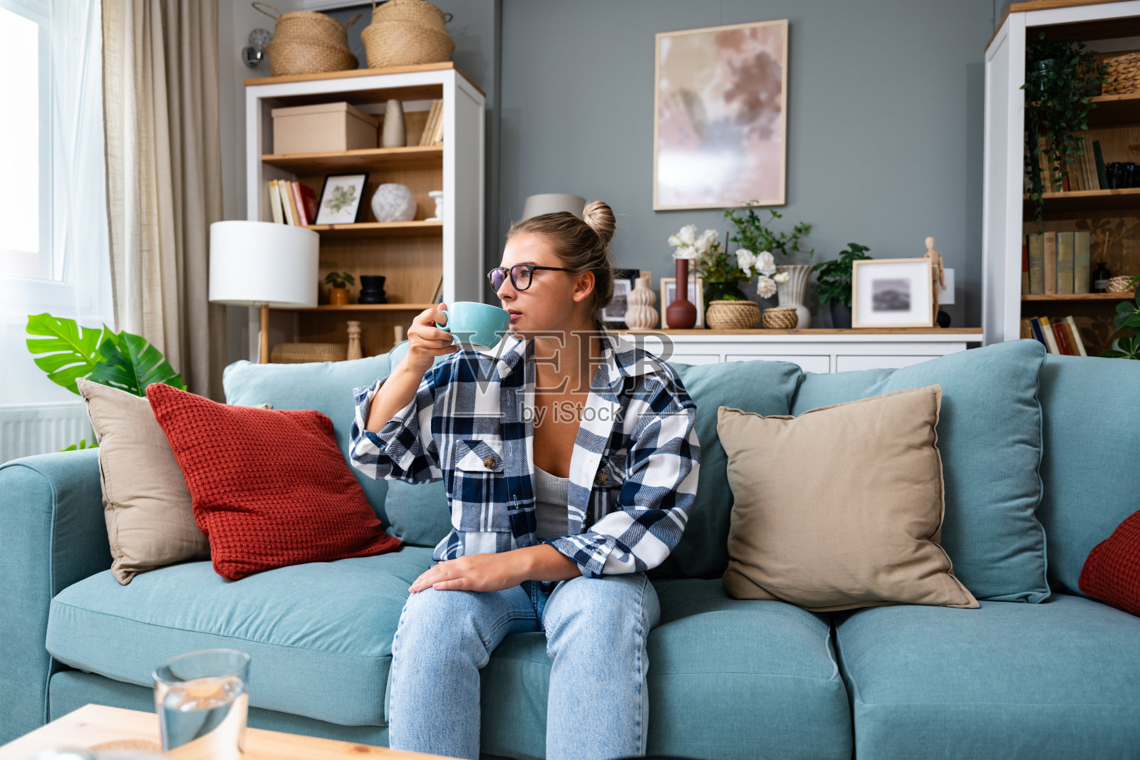 年轻的微笑的女人坐在沙发上，看着喝热茶或咖啡。年轻的黑发女人闲暇时在家里思考。在一个明媚的冬日早晨，快乐的女孩在家里放松。照片摄影图片
