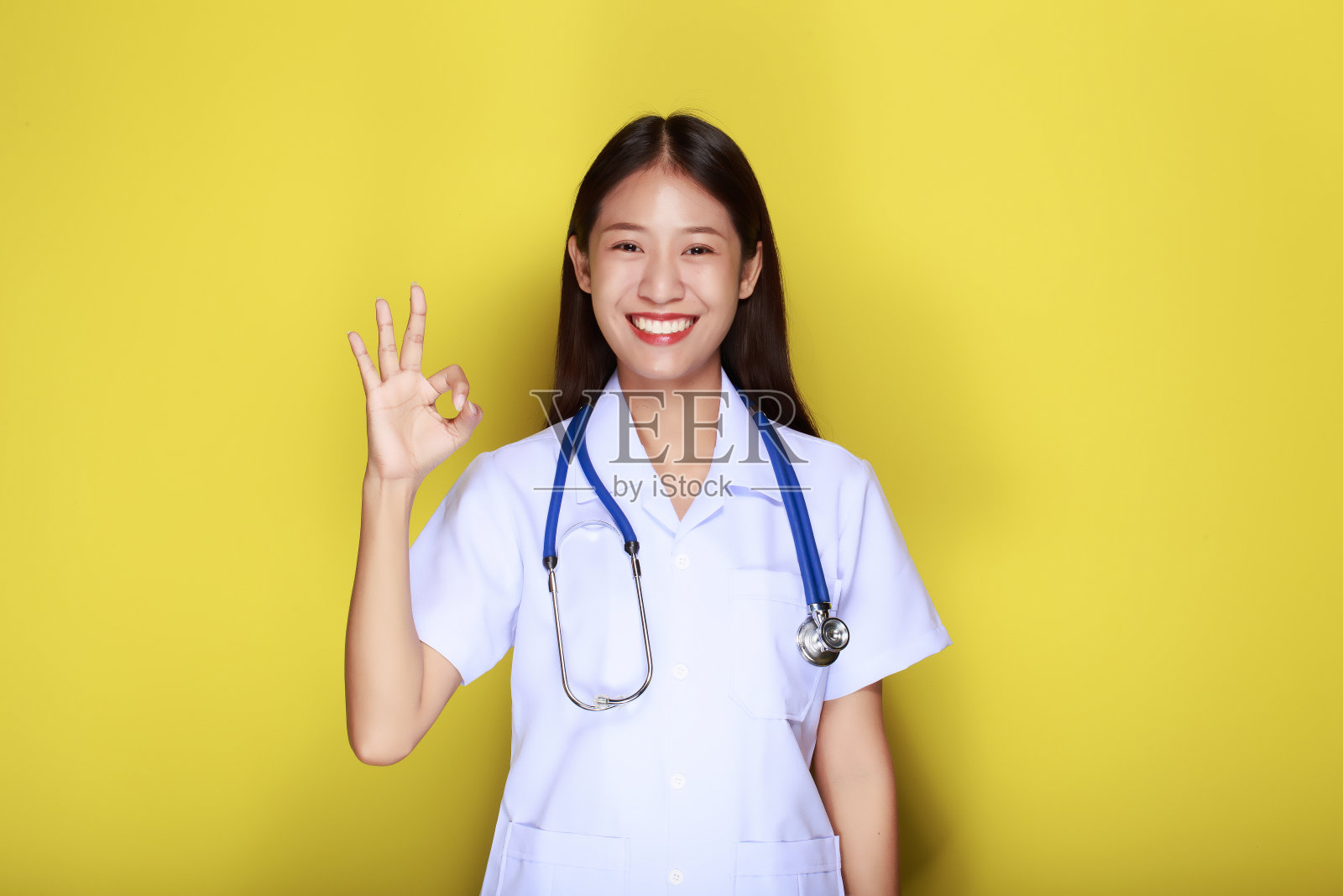 一个美丽的年轻女子在黄色背景的肖像，美丽的亚洲妇女站着愉快地用手做出OK的手势，穿着医生的制服。照片摄影图片