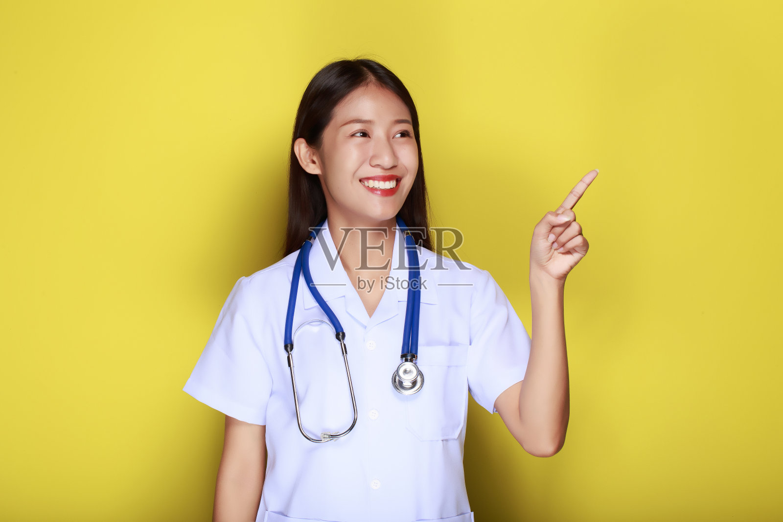 一个美丽的年轻女子在黄色背景的肖像，友好的美丽的女人穿着医生的制服，戴着听诊器指着。照片摄影图片