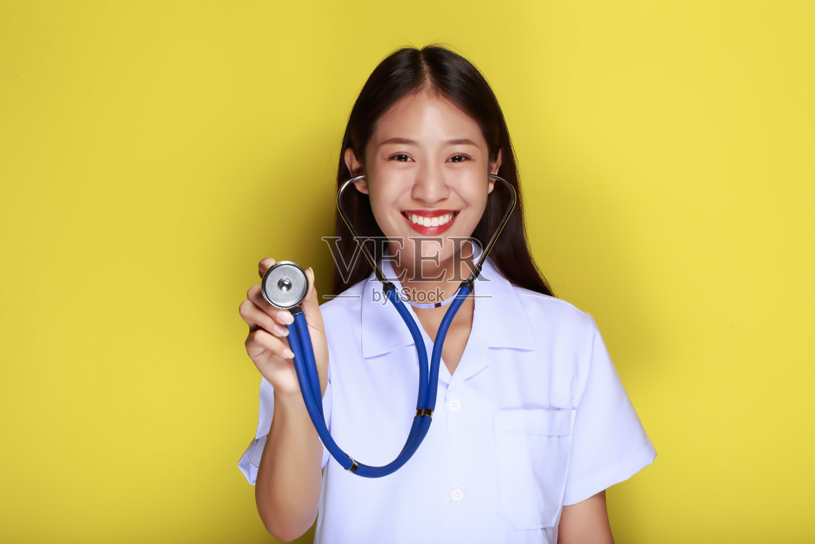 在黄色背景下，一位美丽的年轻女子的肖像，一位穿着医生制服的亚洲妇女露出微笑的表情，一位戴着医用听诊器的年轻女子露出友好的表情。照片摄影图片