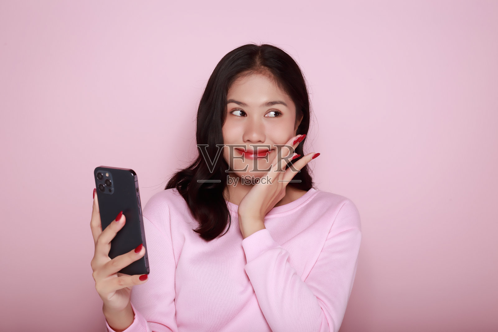 亚洲女子愉快地摆姿势与手机，一个美丽的年轻女子感到愉快和幸福。一个美丽的年轻女子的肖像在浅粉色的背景，照片摄影图片