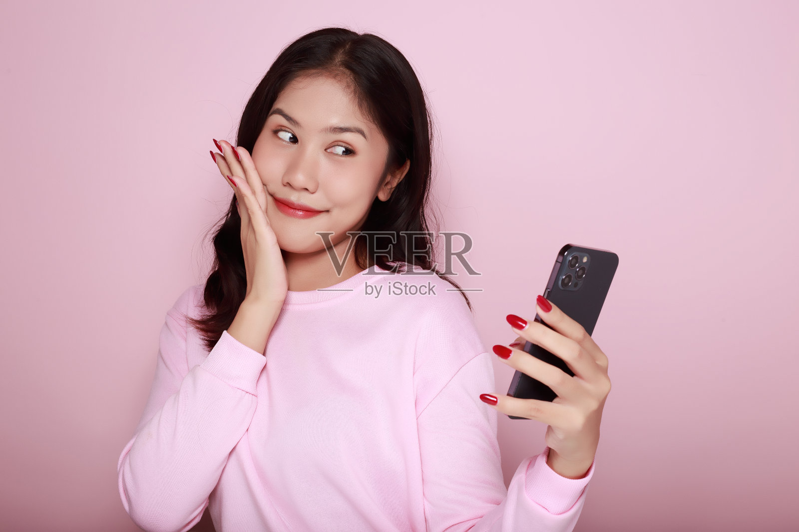 亚洲女子愉快地摆姿势与手机，一个美丽的年轻女子感到愉快和幸福。一个美丽的年轻女子的肖像在浅粉色的背景，照片摄影图片