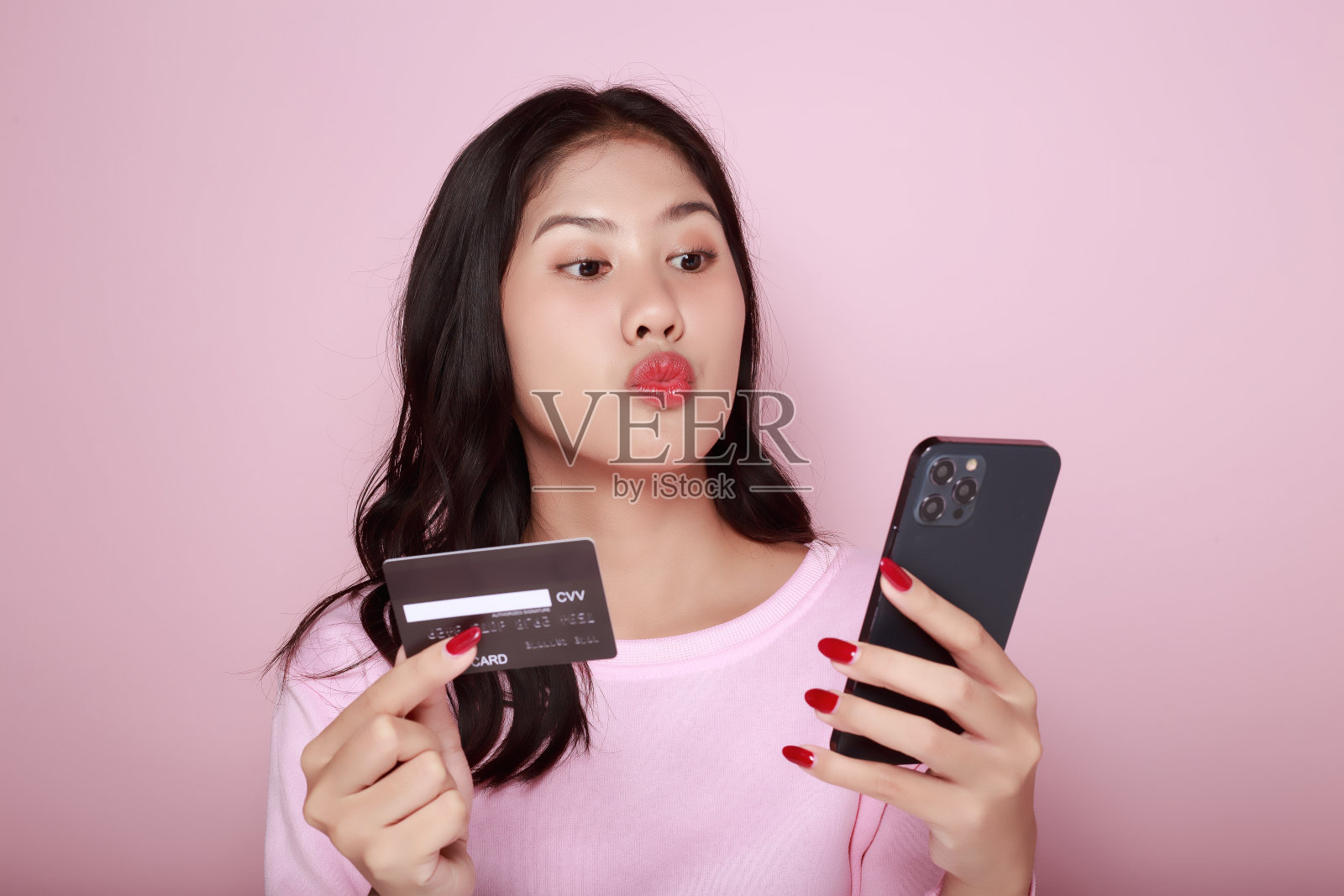 年轻女子手里拿着智能手机和信用卡。一名亚洲女子在浅粉色背景上拿着装备摆姿势。照片摄影图片