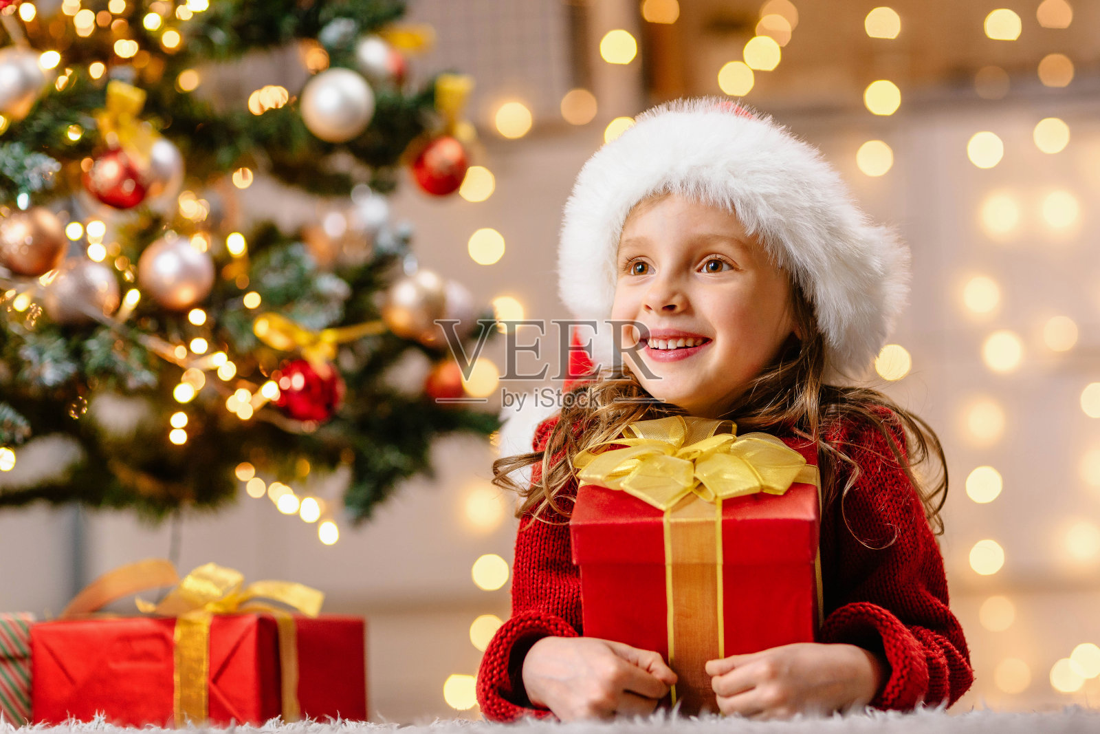 这个快乐的女孩对这份礼物很满意，她望向远方。一个孩子站在一棵装饰好的圣诞树旁照片摄影图片