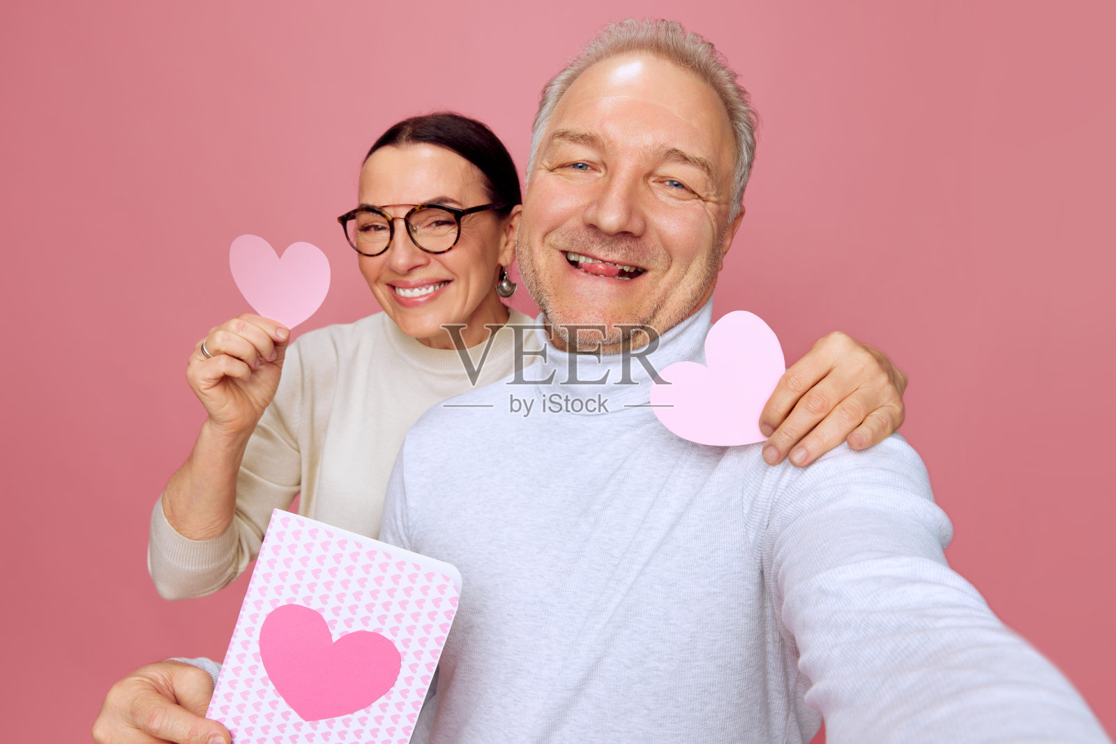 有问题的时间。快乐，微笑的老男人和女人在粉红色的工作室背景下用纸心形明信片拍照。情人节照片摄影图片