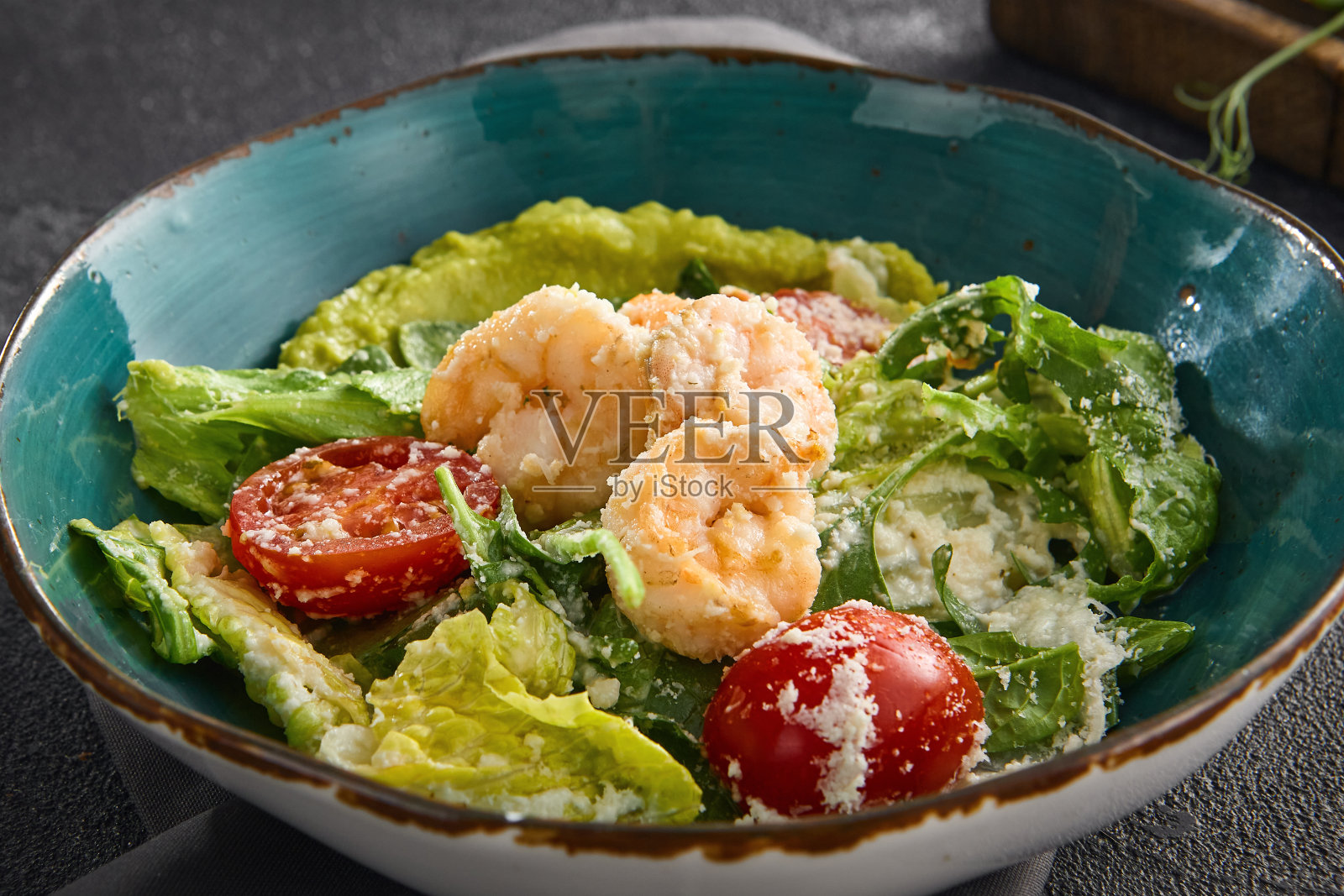 优雅的凯撒虾沙拉装在蓝绿色的碗里，在深色的桌子上，突出了多汁的虾和成熟的圣女果照片摄影图片