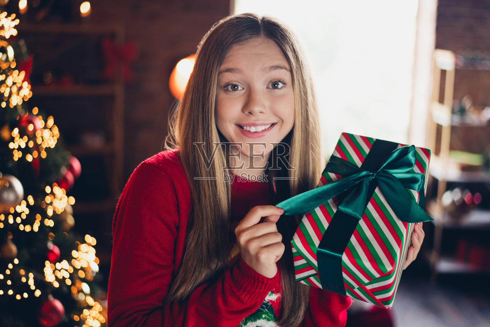可爱的好心情的女学生的照片，金色的发型，穿着红色的毛衣，圣诞节早上在家里打开礼物照片摄影图片