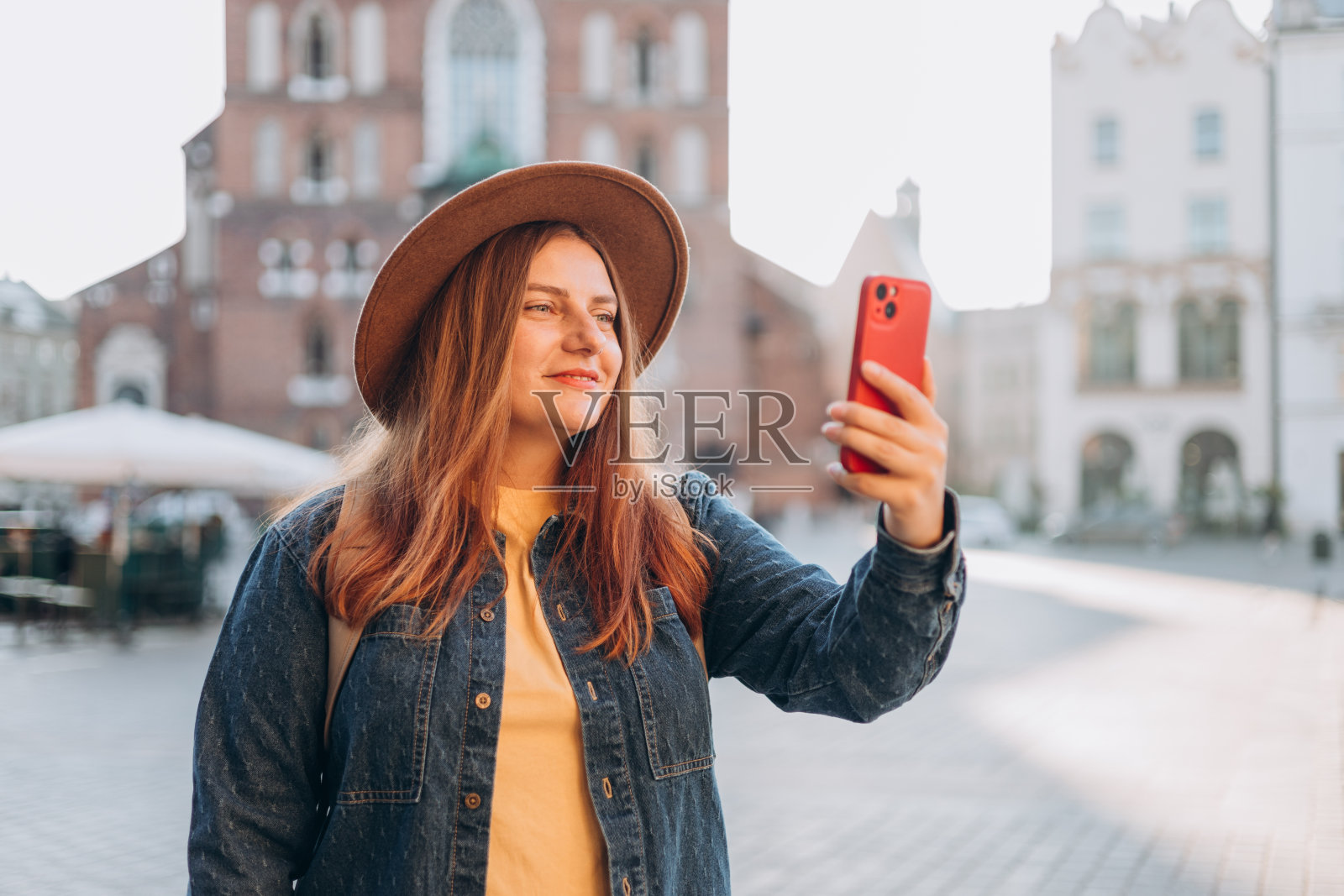 波兰克拉科夫市场广场上著名的圣玛丽大教堂前，戴着帽子的年轻女游客正在自拍。秋天去欧洲旅行。假期的概念照片摄影图片