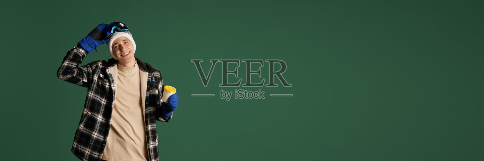 穿着格子夹克，戴着帽子，戴着护目镜的年轻人微笑着站在绿色的工作室背景下，拿着咖啡杯。极限运动照片摄影图片