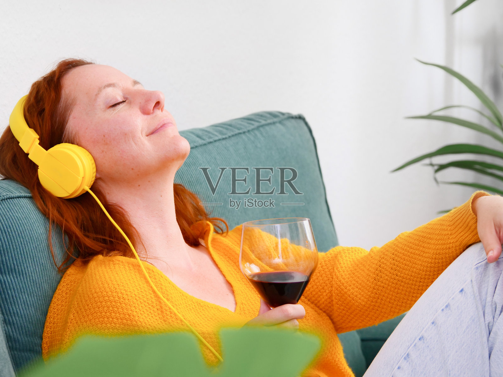 戴着黄色耳机、端着一杯红酒的女人坐在家里的沙发上听音乐或播客。音乐和放松照片摄影图片