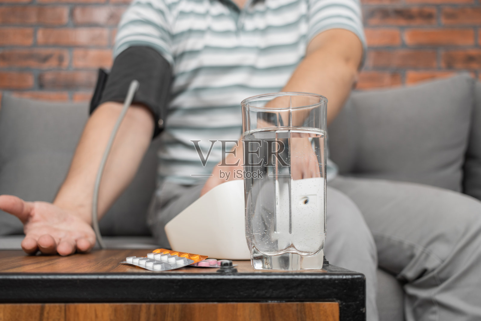 散焦的背景是男人坐在家里的沙发上，用现代数字血压计测量手上的血压，焦点是前景上装有药片的泡罩包装照片摄影图片