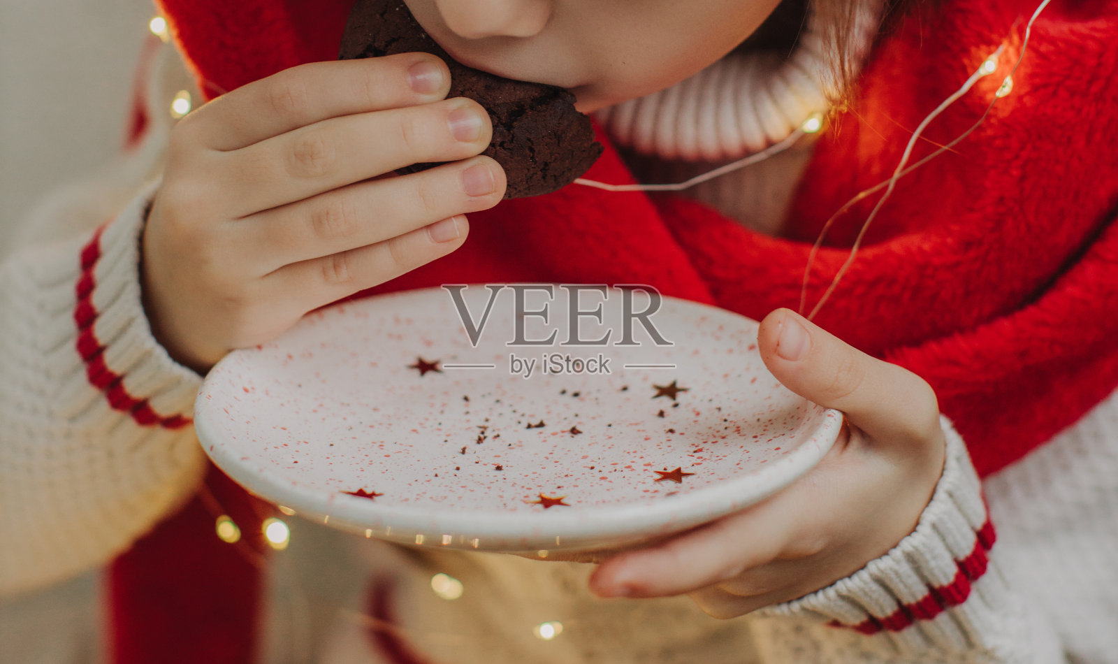一个十几岁女孩的手拿着饼干，背景是圣诞老人的帽子和花环灯。喜庆的新年食物概念。特写照片。照片摄影图片