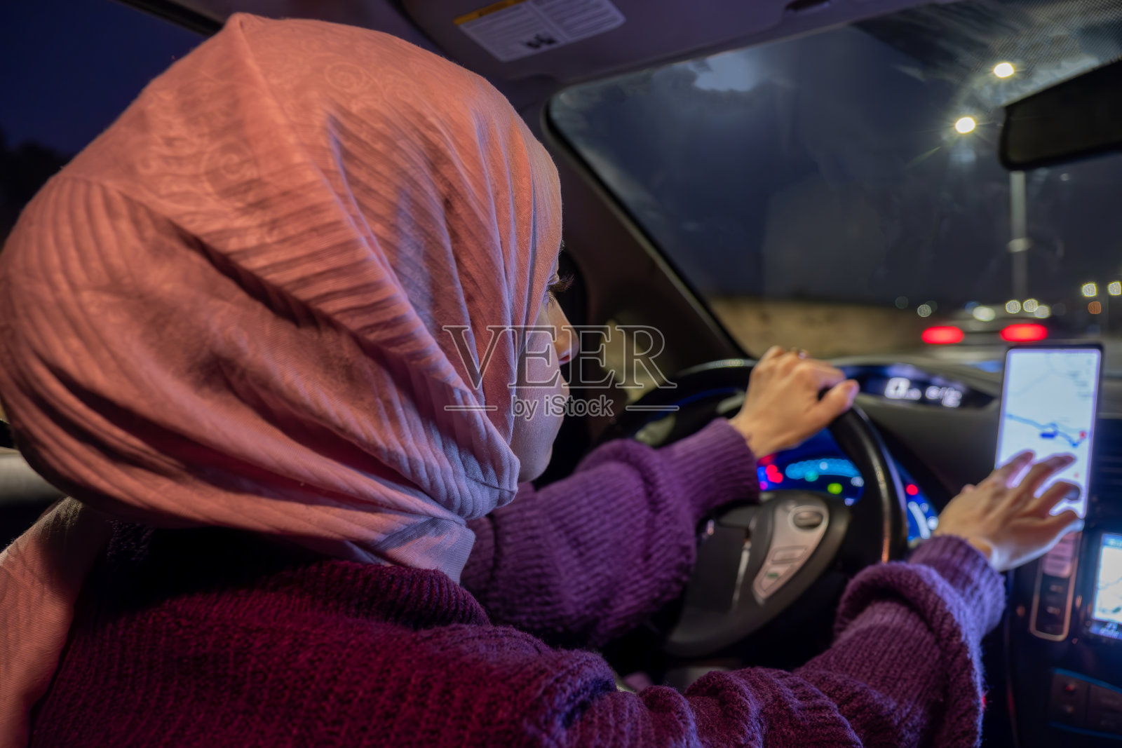 阿拉伯穆斯林妇女晚上开车，穿着冬衣，戴着头巾，系着安全带，自信地开车，通过手机和GPS使用地图照片摄影图片