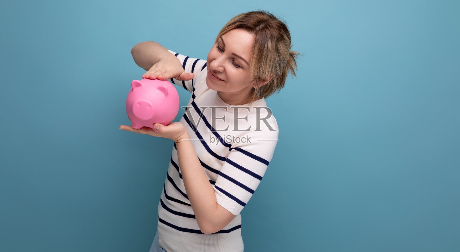 有魅力的年轻女子的横向照片，穿着休闲服装，可爱地抱着一个存钱罐，蓝色背景上有复制空间照片摄影图片