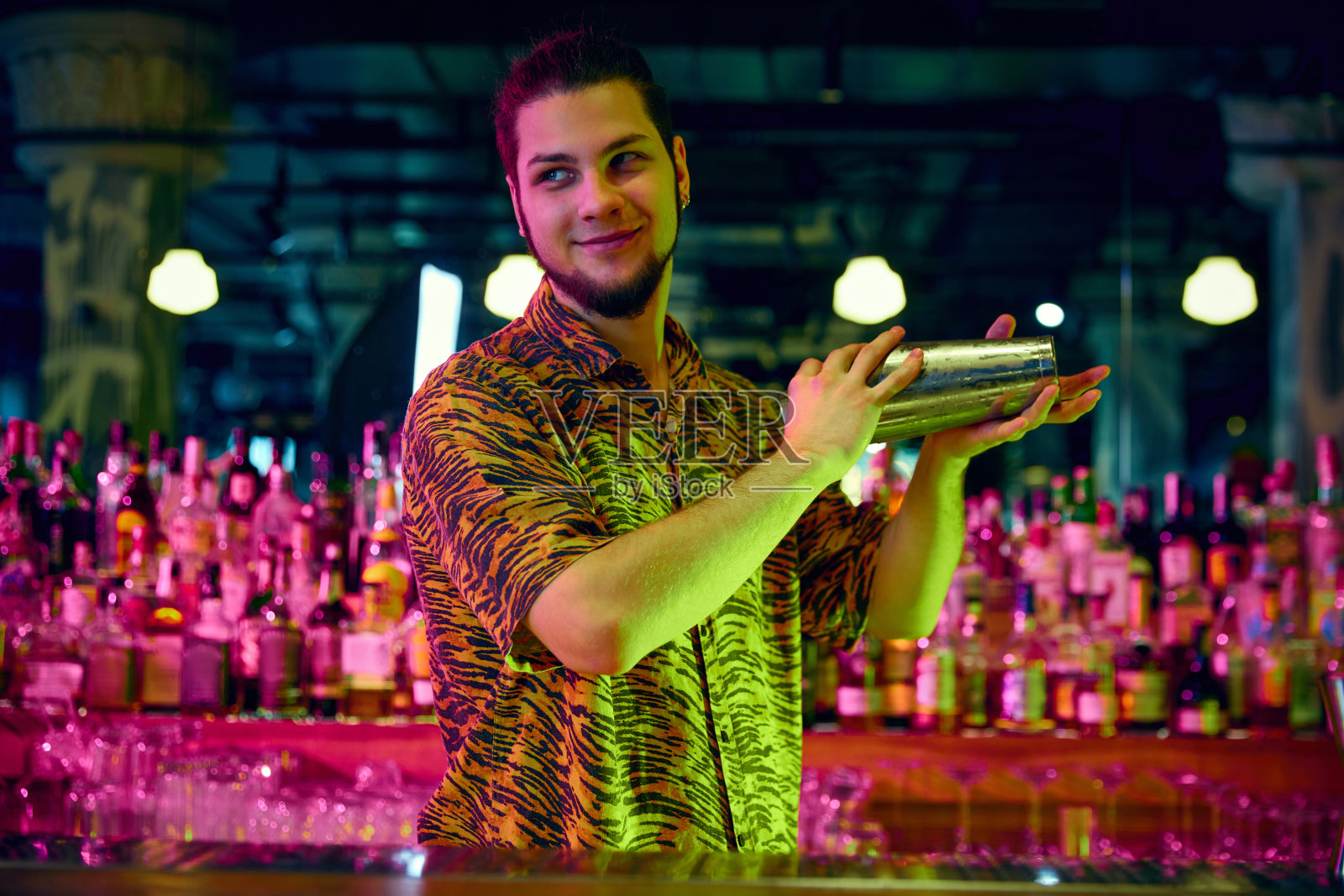 年轻的小伙子，调酒师摇着鸡尾酒摇壶，在现代设计和霓虹灯的酒吧里调制甜美可口的鸡尾酒。照片摄影图片