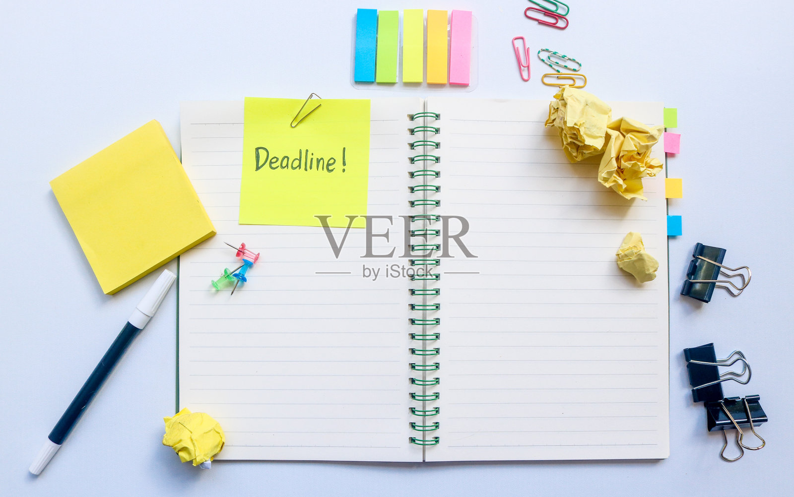 最后期限这个词写在笔记本顶部的黄色便签上，与其他物品(如笔、回形针、活页夹、皱巴巴的纸、便签)隔离在白色的背景上照片摄影图片